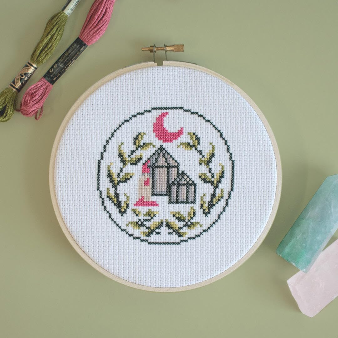 Cross-stitched design with crystals and moon on white fabric in an embroidery hoop, with embroidery threads and tools on a green background.