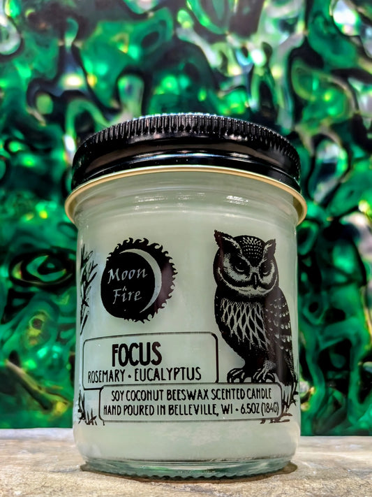 Candle jar labeled 'Focus' with owl design and 'Moon Fire' branding on a green abstract background